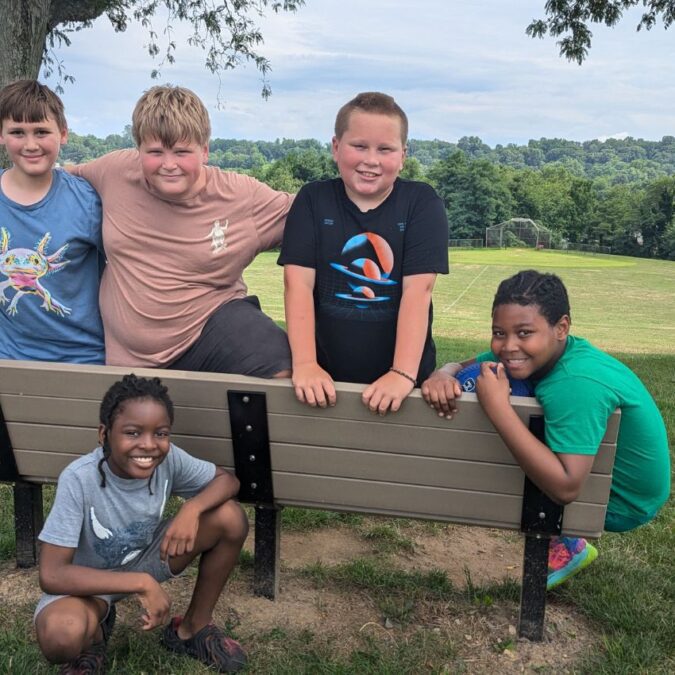 A group of boys on a bench outside near a soccer field smiling for the camera.