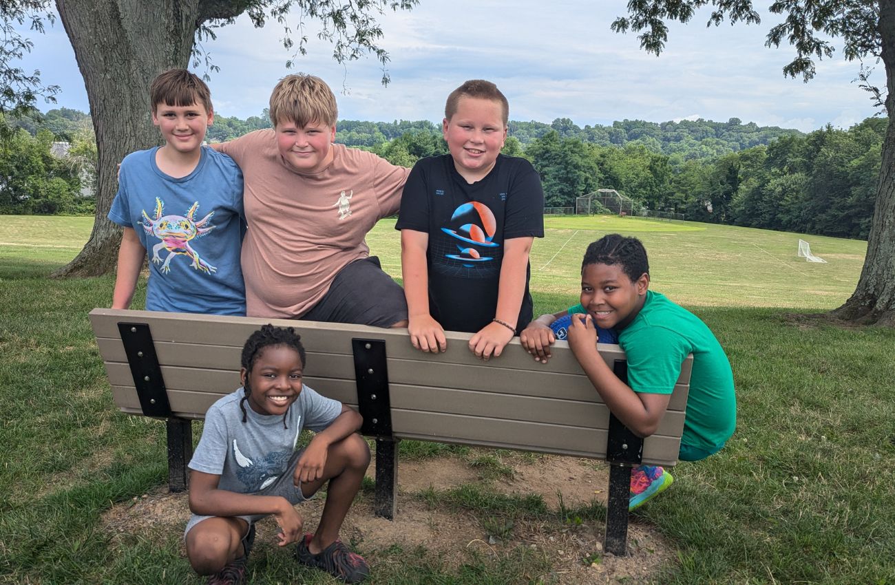 A group of boys on a bench outside near a soccer field smiling for the camera.
