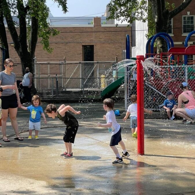Little kids with parents nearby cool off under a red sprayer at the sprayground in Starr Garden, a Center City playground.
