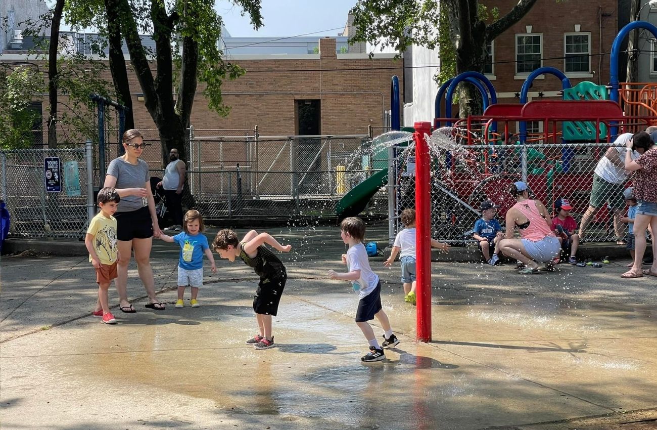 Little kids with parents nearby cool off under a red sprayer at the sprayground in Starr Garden, a Center City playground.