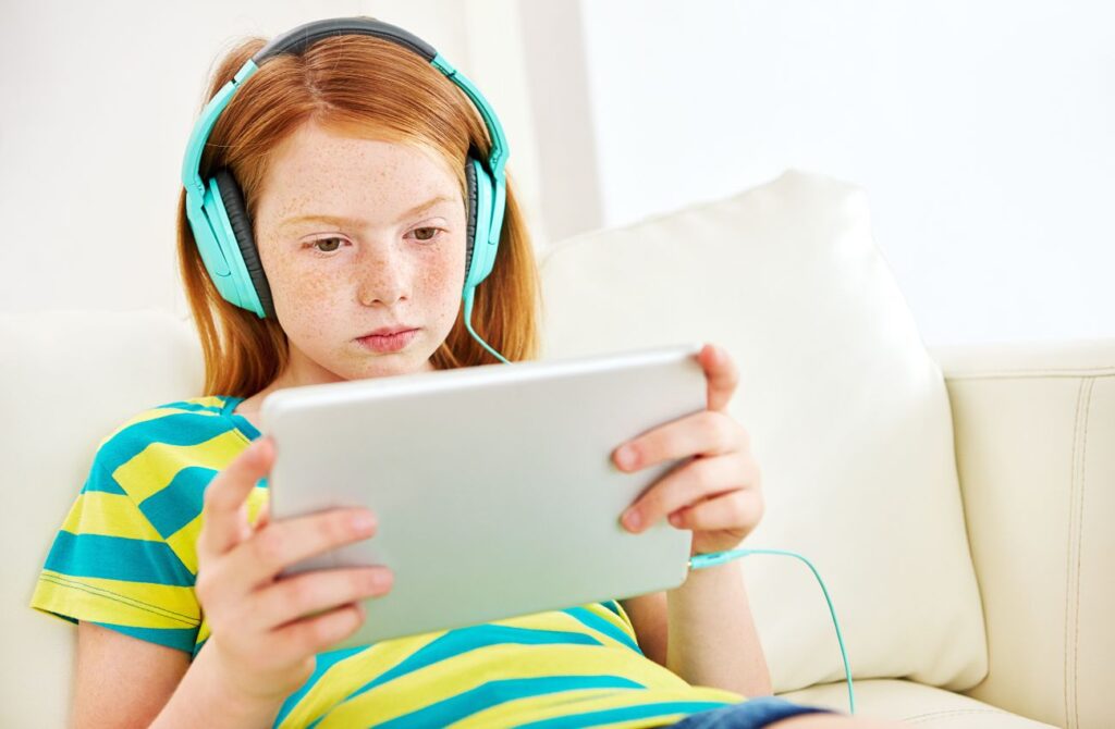 An elementary school-aged girl wears headphones while looking at a tablet device.