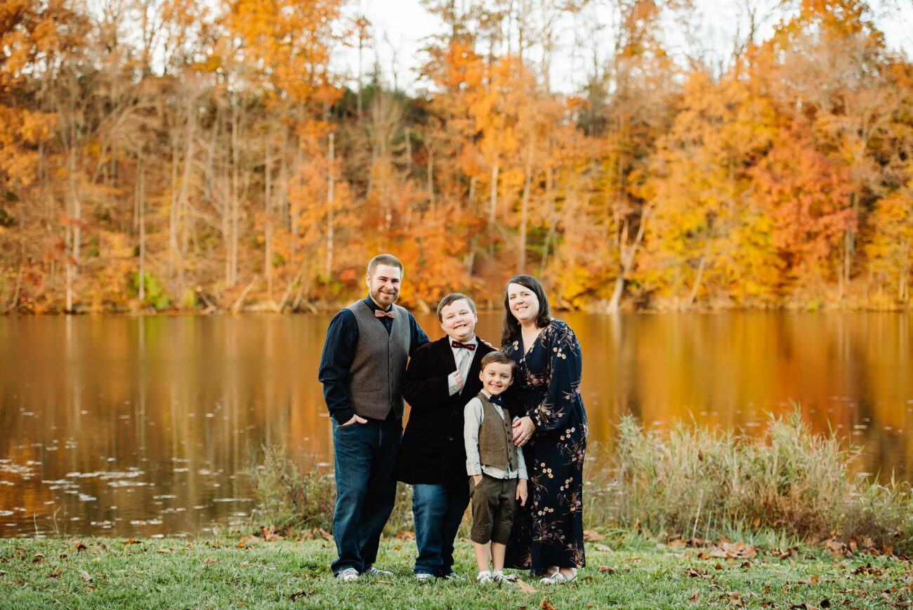 The Thomas Family with two young boys in front of a lake with fall foliage behind them