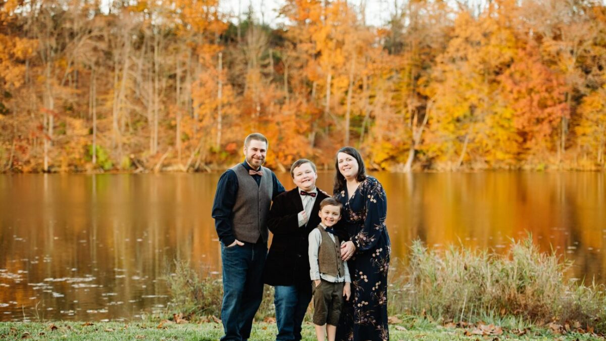 The Thomas Family with two young boys in front of a lake with fall foliage behind them