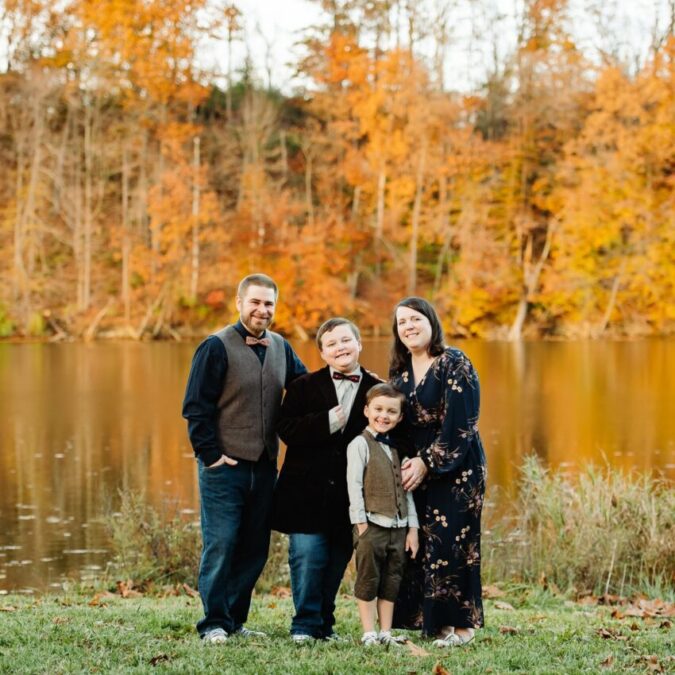 The Thomas Family with two young boys in front of a lake with fall foliage behind them