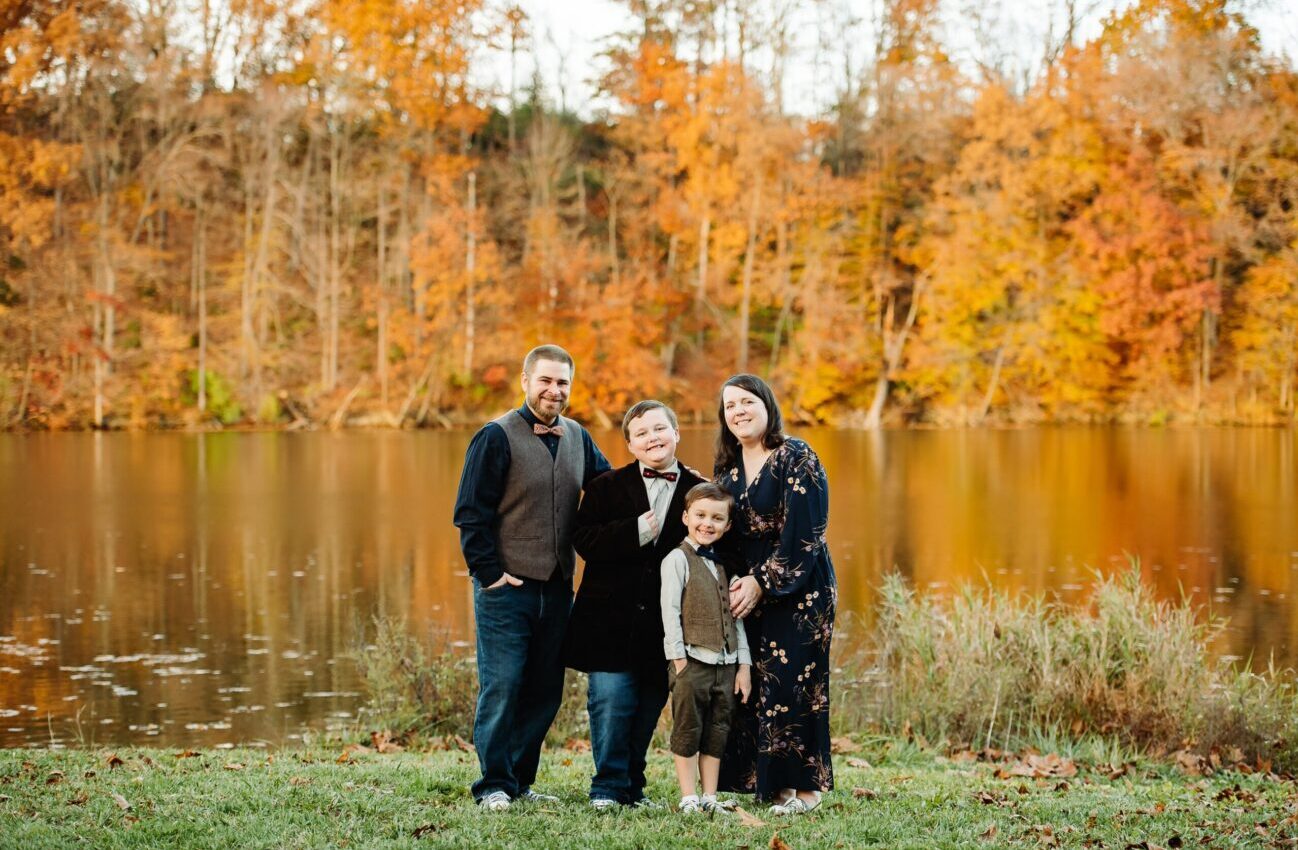The Thomas Family with two young boys in front of a lake with fall foliage behind them
