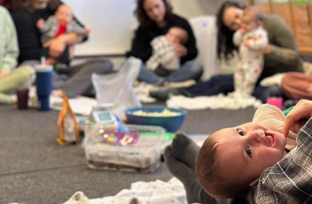 A parent holds a smiling baby in a circle of other parents at Nesting House's new baby meetup.