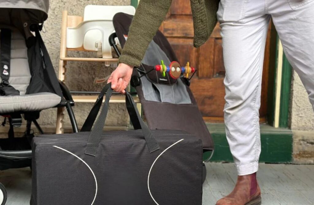 A hand holds a pack and play bag in front of other gently used baby gear.