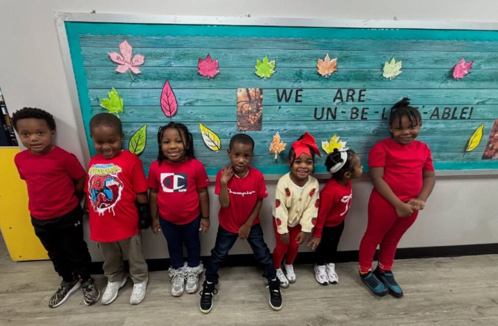 Seven pre-K boys and girls wearing red line up along a blue bulletin board in class. 
