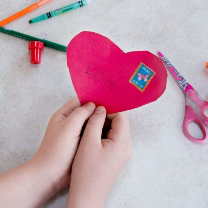A child's hands hold a red paper heart that says Love for Valentine's Day.