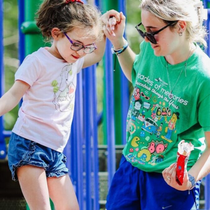 A counselor at JCC Camps in Medford supports a camper with disabilities on play equipment.