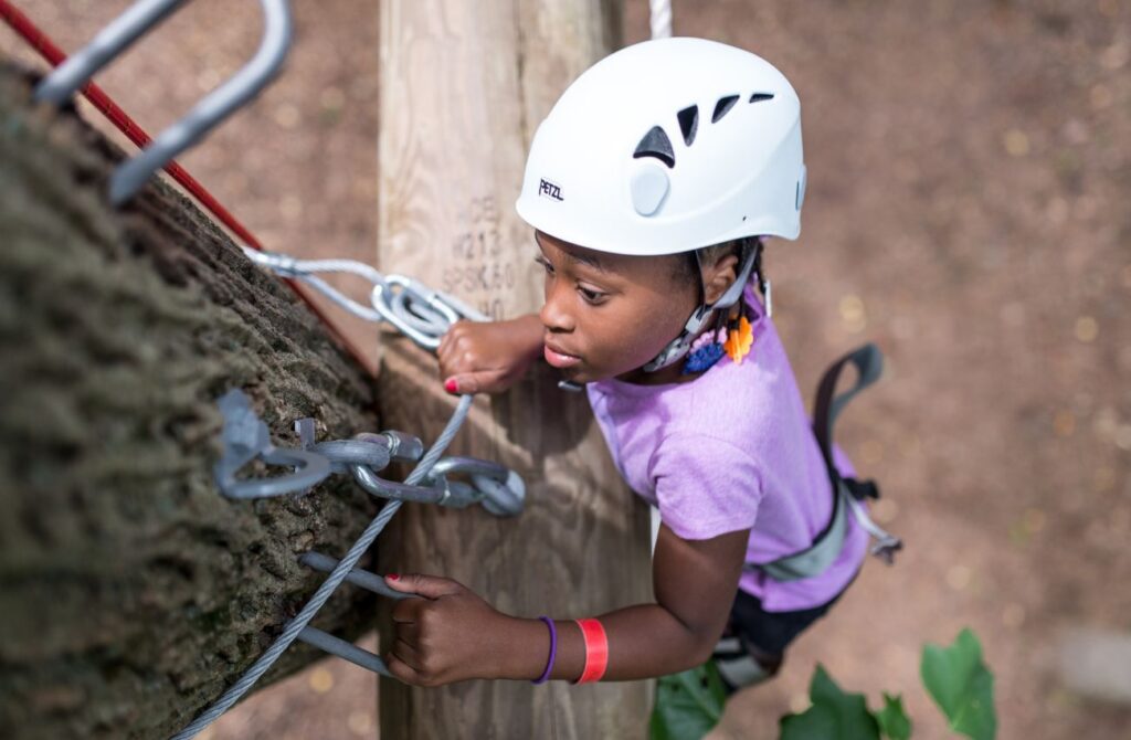 A camper in climbing gear and helmet climbs a tree at Camp Speers YMCA. 