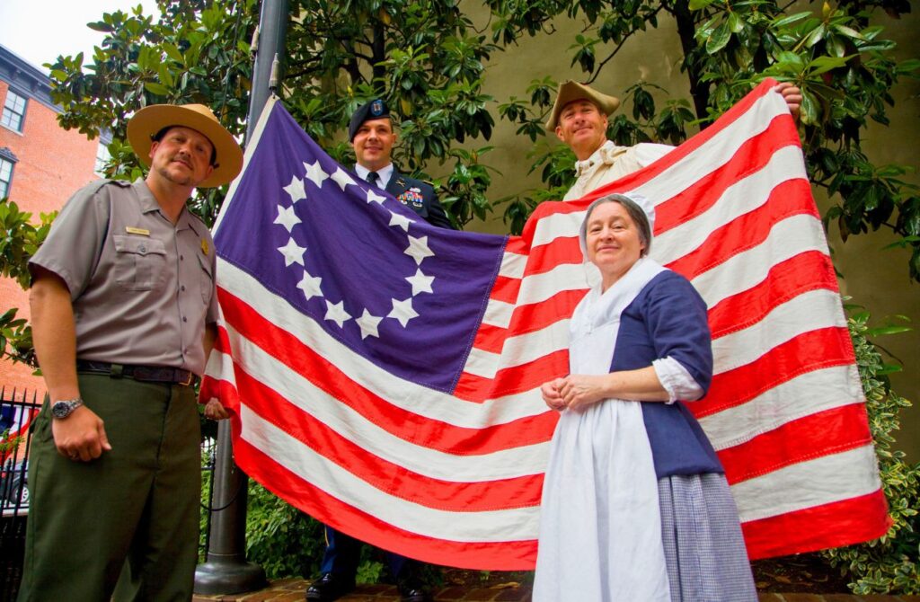 A costumed Betsy Ross and colonial-era man pose with a US soldier and park ranger in front of the colonial American flag with 13 stars. 
