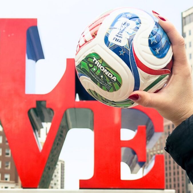 A hand holds a FIFA World Cup 2026 mini soccer ball in front of the LOVE sculpture in Philadelphia.