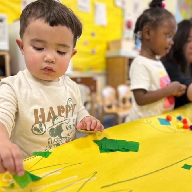 A boy sticks paper cutouts on a bright yellow table pattern at a free Philadelphia Pre-K class.